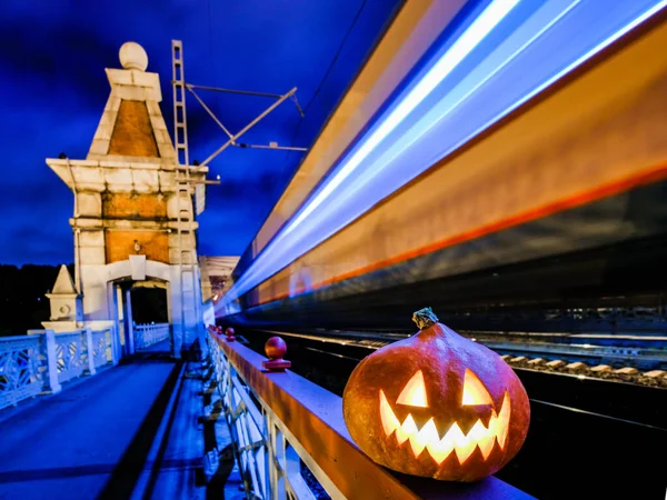 Halloween pumpkin with a glowing grimace at night on the railway bridge. In the background there are rails and blurry streaks of lights from a speeding train. Mystical Halloween in the city.