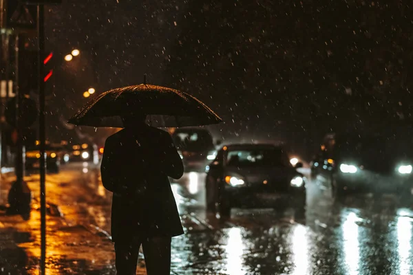 Silhouet van een man met een paraplu in de achtergrondverlichting van koplampen 's nachts tijdens een regenbui. Zware regenval in de stad — Stockfoto