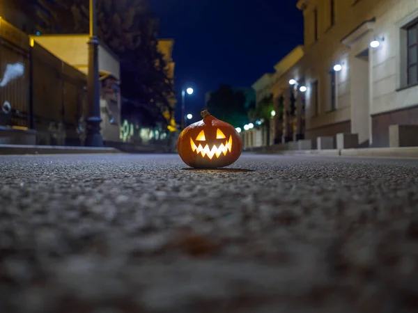 Halloween pumpkin on the asphalt on a deserted city street at night. Blurry city lights and old multi-story buildings. The decor of the night city in a gloomy festive Halloween style. Copy space.