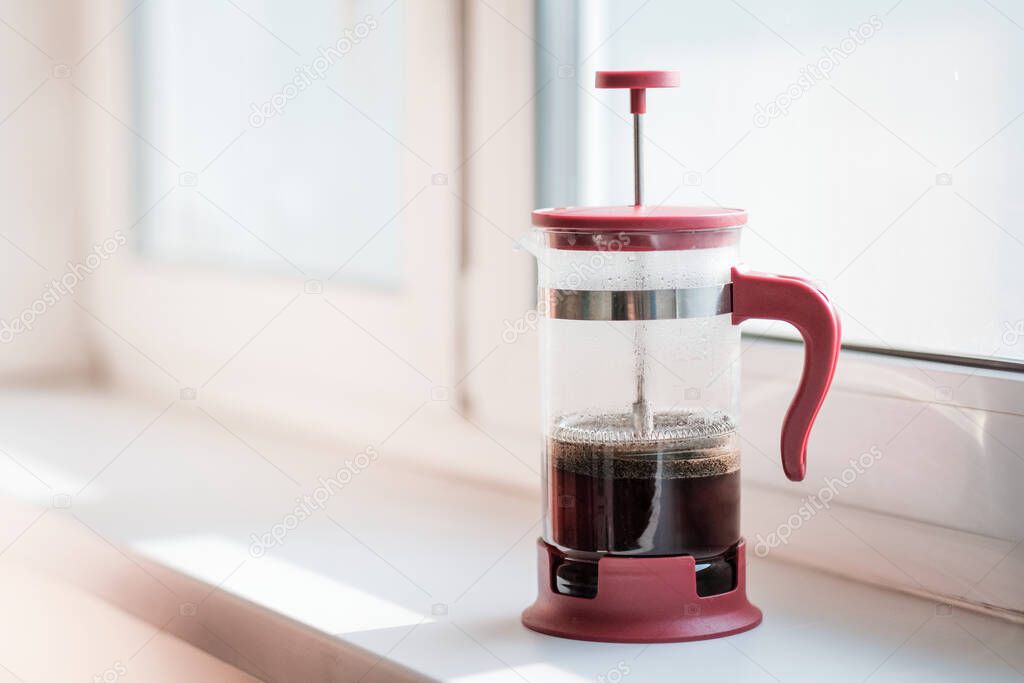 Glass Frenchpress on the windowsill in the rays of bright sunlight. The process of brewing ground black coffee. A fragrant drink made from roasted grains gives pleasure and vigor. High quality photo.