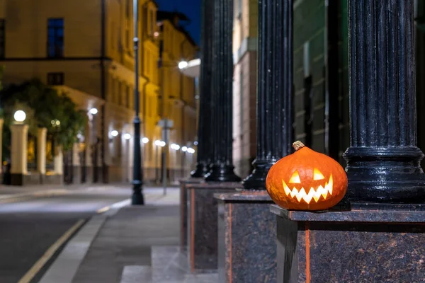 Halloween pumpkin on a deserted city street at night. Blurry colored city lights and old high-rise buildings. The decor of the night city in a festive Halloween theme. Copy space.