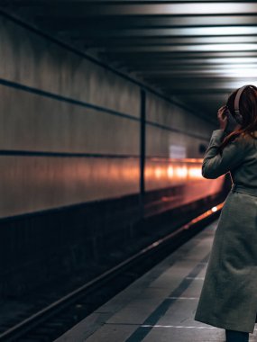 Seçici odaklanma. Kafasında kulaklık olan gri montlu bir kız. Kız müzik dinliyor. Trenin arka farlarının ışığı karanlık bir tüneldeki bir kızın siluetini aydınlatır.