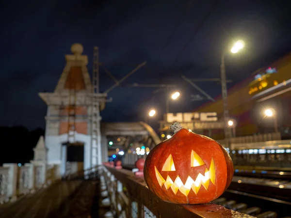 Halloween pumpkin with a glowing grimace at night on the railway bridge. In the background there are rails and blurry streaks of lights. Mystical picture of Halloween in the city