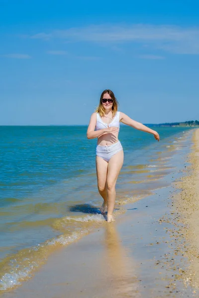 Ein junges Mädchen läuft am Sandstrand entlang der Brandung. Spritzer Meerwasser. Das Mädchen trägt einen weißen Badeanzug und eine Sonnenbrille. Freiheit und Sorglosigkeit. Fitness im Freien. Sommersonniger Tag — Stockfoto