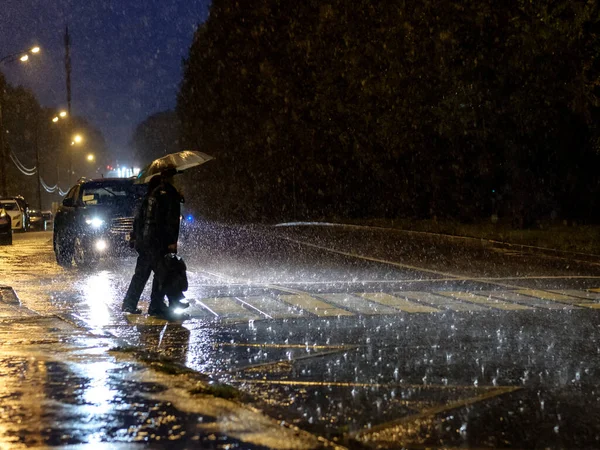 Utsikt över en fotgängare korsning i staden på natten under en tung skyfall. Silhuetter av personer med paraplyer. — Stockfoto