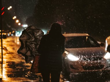 Gece yarısı arabaların farlarının ışığında bir kadının silueti. Kadın taksiye gider ve şemsiyeyi katlar.