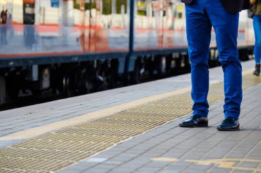 Gelen trenin arka planında parlak mavi pantolonlu ve siyah deri ayakkabılı bir adamın bacakları.