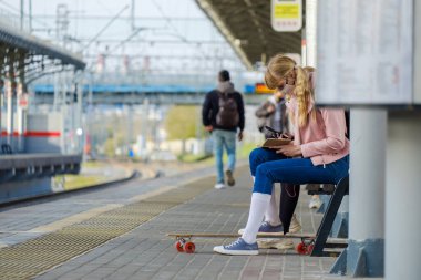 Moskova. - Rusya. 4 Ekim 2020. Sarı saçlı, koruyucu maskeli bir genç kız tren istasyonundaki bankta oturuyor.