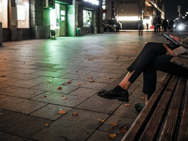 Beautiful elegant womens legs in black glossy loafers. A woman sits on a bench on a night street in the city in autumn