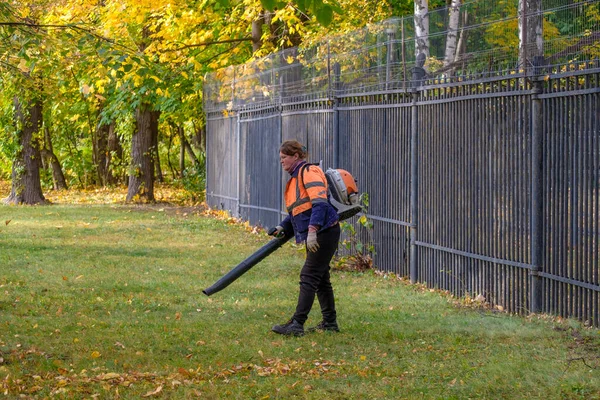 Moskva. Ryssland. 11 oktober 2020: En kvinnlig bruksarbetare använder en fläkt för att ta bort fallna löv på en gräsmatta i en park. — Stockfoto