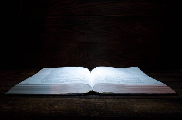 The big book of the Bible lies on a wooden table. In the dark. A light shines on the book from above.