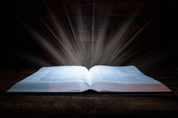 The big book of the Bible lies on a wooden table. In the dark. A light shines on the book from above. Light comes out of the book