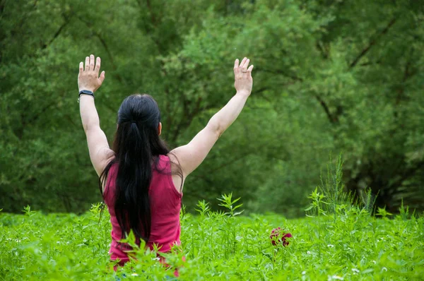 a woman in a burgundy vest stands among tall green grass and plants with arms raised. Summer in the garden. Tall trees