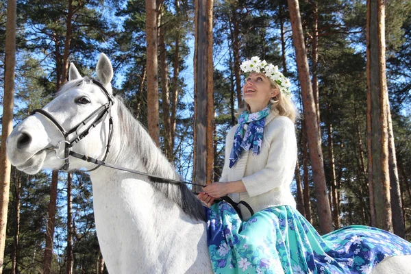 Beautiful young blond woman with flower crown on white horse in sunny winter day in the forest as a symbol of coming spring