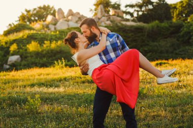 Onun güzel kız arkadaşı kollarında taşıyan ve çim alanda öpüşme genç adam bir kadeh. Doğada bir yaz günü zevk çift.