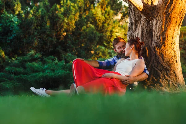 美しいカップルが公園で市内外で休み お互いに庭の一見に大きな木の近くに座って 笑顔します 愛の物語 — ストック写真