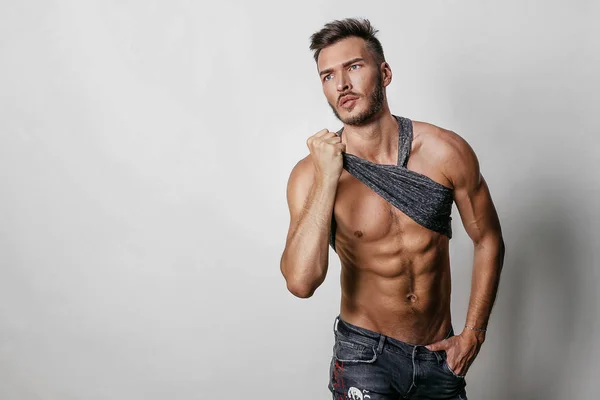 Kas modeli spor genç adam Mavi Jeans kaldırdı onun t-shirt ve onun basın gri bir arka plan üzerinde gösterilen El. Sportif sağlıklı güçlü muscleguy moda portre. Seksi gövde.