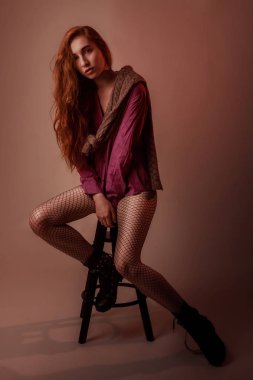 Omuz ve sandalye sıcak tonları üzerinde oturan siyah File külotlu çorap, kazak ve gömlek kırmızı uzun saçlı kadın portresi Studio moda ve stil kavramı