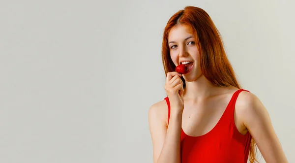 ポーズとスタジオでイチゴを食べる赤い水着でセクシーなスリムな女性 完全な長さの長いウェーブのかかった赤い髪の美しい少女ファッションの肖像画 灰色の背景をコピーします — ストック写真