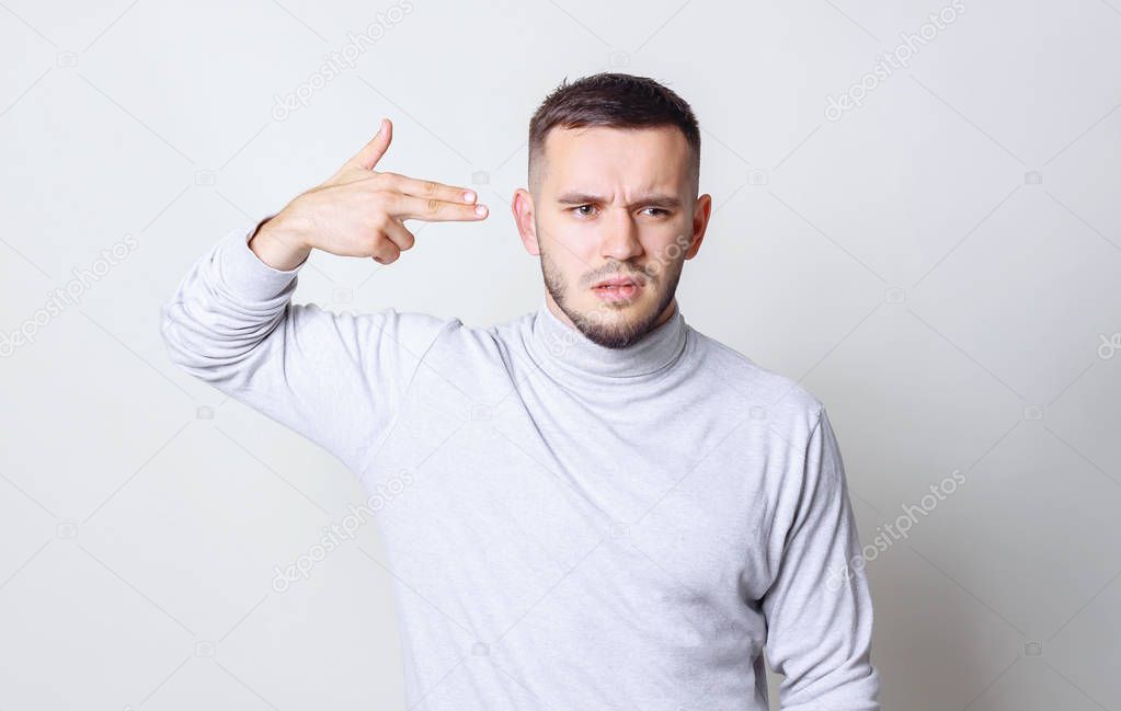 Young man committing suicide with finger gun gesture, shooting himself in head making finger pistol sign, man in white turtleneck on grey background copy space