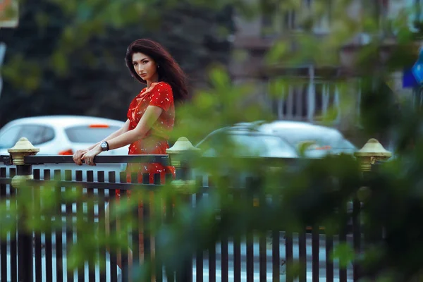 橋の上フェンス近くに美しい日焼け少女が立っています 自然の背景をぼかした写真の素敵な女の子が 赤いドレスを着たかわいい女の子 美しい若い女性の日常生活 — ストック写真