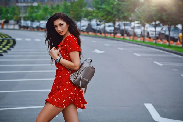 立っている若いブルネットの少女 赤背景の場所を駐車場街に肩に身に着けているバックパックとショート ドレスでポーズ — ストック写真