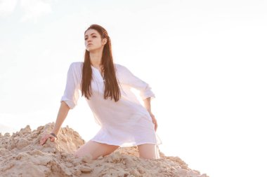 Şirin esmer kadın beyaz bir bluz ve çıplak bacak kum üzerine oturma, dinlenme ve çöl güneşli gün günbatımı gökyüzü üzerinde poz poz zarif