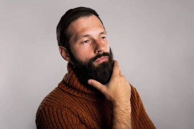 Üzerinde bir şey düşünüyorum yakışıklı bir adam portresi. Kendini uzun kalın bir sakal okşayarak gri arka plan üzerinde izole sıcak örme kazak giymiş.