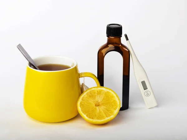 A yellow cup of tea with lemon, a thermometer and bottle cough syrup for treatment Cold And Flu. Prevention of colds and flu on grey background