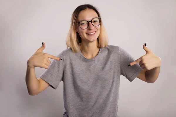 The smile girl points fingers at herself with both hands. Charming young girl in a gray T-shirt and big eyeglasses on a gray background.
