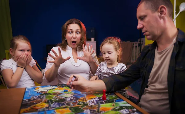 Board game concept. The family is having fun playing together at home in a board game. Board game field, many figures, coins and sandglass. Four people play, holding cards joyful emotions on the face.