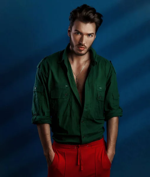 Fashion portrait of young man in green shirt and red pants poses over dark blue wall with contrast light