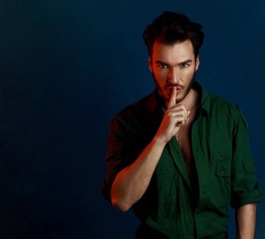 Adam yeşil gömlek ve stil saç kesimi pozlar kırmızı kontrast renk ışık ile karanlık mavi duvarın üzerinden moda karanlık tarzı portresi susturmak için veya sessiz olmak isteyen yakışıklı erkek shhh hareketi ile kapatmak