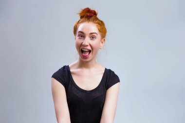 Kızıl saçlı hayran kadın modeli dinleme cihazı gözlerle bakarak ve yaygın olarak gri arka plan üzerine ağzını açtı siyah gömlek giymiş düğüm bağladılar