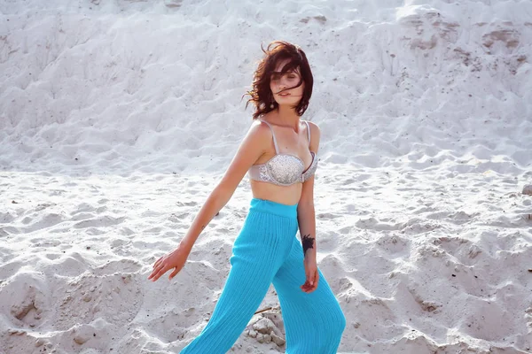 Portré, fiatal, gyönyörű nő pózol a homokos strand, vagy a sivatagban. Modell, elegáns kék nadrágot és a melltartó viselése. Naplemente fény. Női divat fogalmát. — Stock Fotó