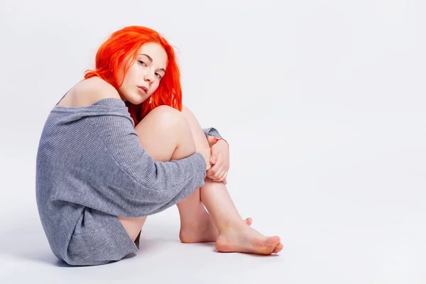 Retrato de moda de la hermosa pelirroja adolescente vestida con suéter de punto piernas desnudas mirando por encima del hombro mientras posa sobre fondo gris sentado en el suelo, espacio de copia . — Foto de Stock