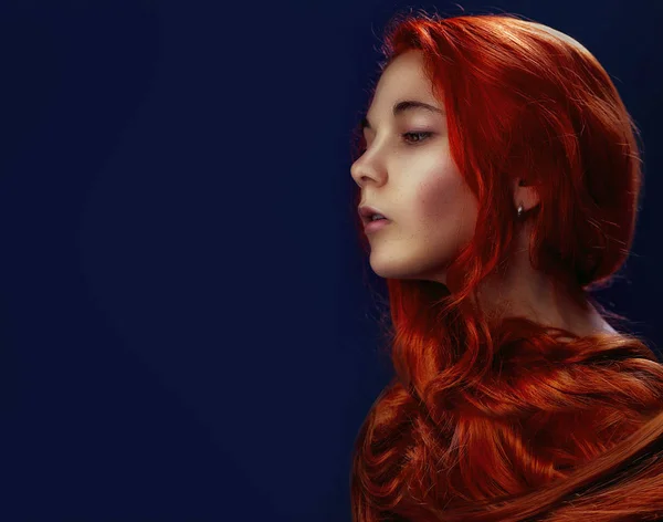 美丽的模型女孩与长卷曲的红头发。时尚女性肖像造型发型卷发侧视图。深蓝色背景上的波浪和闪亮的漩涡 — 图库照片