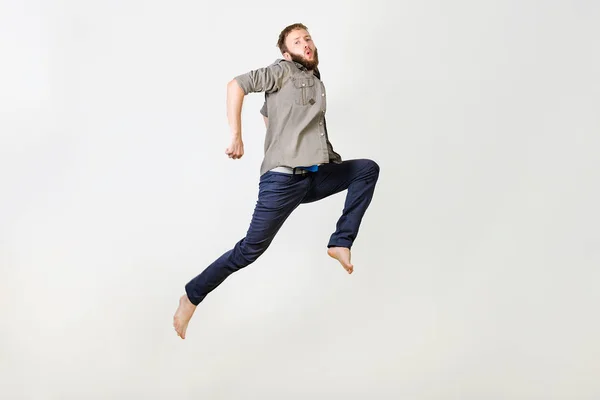 Dlouhá fotka vousatý muž v příležitostné košili a džínách, které běží nebo skáčou ve vzduchu, vytváří legrační obličeje přes šedé pozadí — Stock fotografie