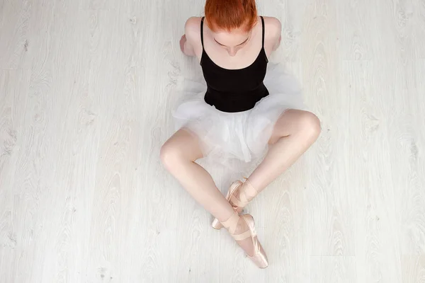 Niza bailarina pelirroja se sienta en el suelo de madera blanca Ella lleva un traje de baile ligero y un tutú de melocotón. Foto de vista superior. Horizontal. Copiar espacio —  Fotos de Stock