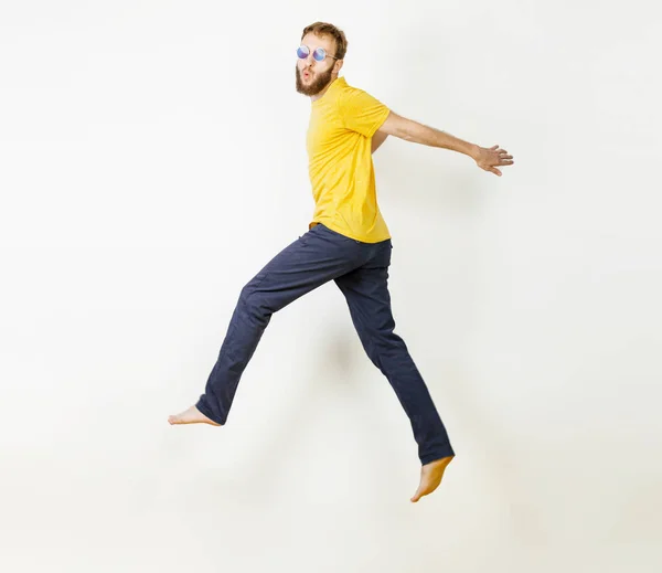 Fotografie muže s plnovousem v příležitostných žlutých Trikách a džínách, které běží nebo skáčou ve vzduchu, vytváří legrační obličeje přes šedé pozadí — Stock fotografie