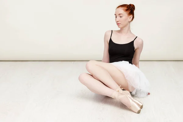 Милая рыжеволосая балерина сидит на полу из белого дерева она носит легкую танцевальную одежду и персиковую пачку. Копирование пространства — стоковое фото