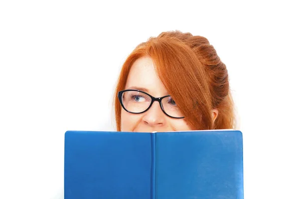 Närbild porträtt av en ung rödhårig kvinna i glasögon gömmer sig bakom en öppen bok isolerad över grå vägg bakgrund — Stockfoto