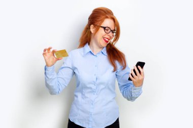 Ayakta ve gri arka plan kopyalama alanı üzerinde izole kredi kartı tutarak cep telefonu bakarak heyecanlı bir kızıl kız Portresi