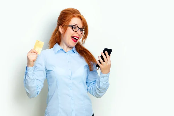 Porträtt av en upphetsad rödhårig flicka tittar på mobiltelefon medan stående och hålla kreditkort isolerade över grå bakgrund kopiera utrymme — Stockfoto