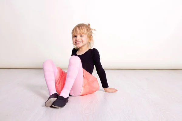 Uma dançarina de tutu cor-de-rosa. Criança pré-escolar sentada no chão de madeira. Bonito criança dançando balé clássico no estúdio branco. As crianças dançam. Jovem dançarina numa aula. Copiar espaço para o seu texto — Fotografia de Stock
