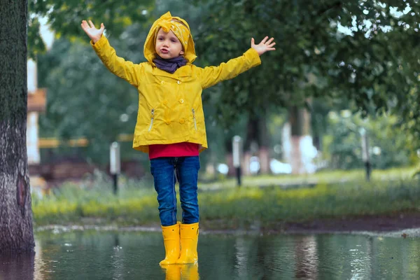 Do not be sad. child girl in yellow raincoat and rubber boots in puddle on a parking, autumn walk. Childhood happiness concept