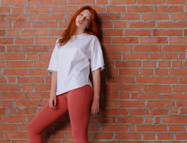 Moda tarzı stüdyo portresi. Güzel, kızıl saçlı, taytlı, beyaz tişörtlü bir kız. Duvarın yanında duran ve poz veren bir model.