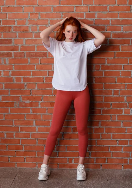Fashion style studio portrait of beautiful red-haired young girl in leggings pants and white t-shirt. Model standing and posing beside brick wall