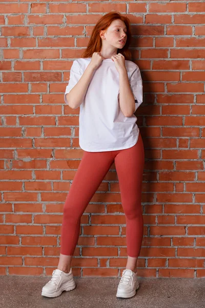 レギンスパンツと白のTシャツで美しい赤髪の若い女の子のファッションスタイルのスタジオの肖像画 レンガの壁の横に立ってポーズモデル — ストック写真