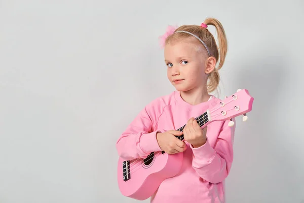 可愛いですCaucasian女の子4 5年古いですピンクスウェットシャツあります遊んでいますザUkulelに対してグレースタジオ背景 — ストック写真